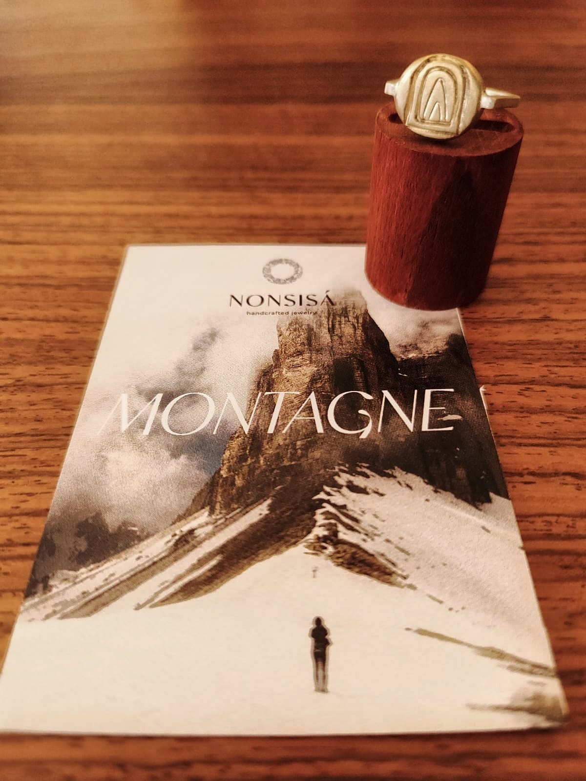 A gold-toned nonsisa ring made of bronze and silver with a stylized mountain emblem, displayed on a dark wooden stand next to its printed packaging card featuring a snowy mountain landscape and the word 'MONTAGNE'.
