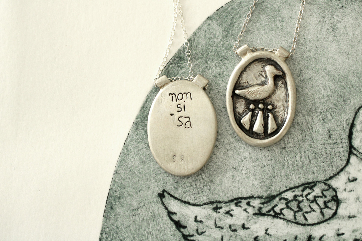 2 Silver necklaces with a circular pendant, left one with the brand name engraved in the back and right one featuring a duck and celtic design on a textured surface.