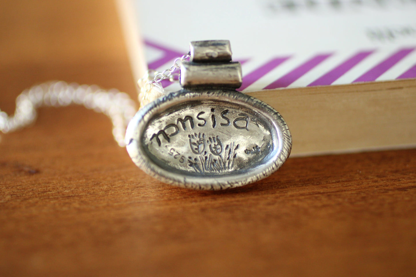 SIlver pendant with a silver chain with an oval shape with the brand name and grain design engraved on the back on a wooden background
