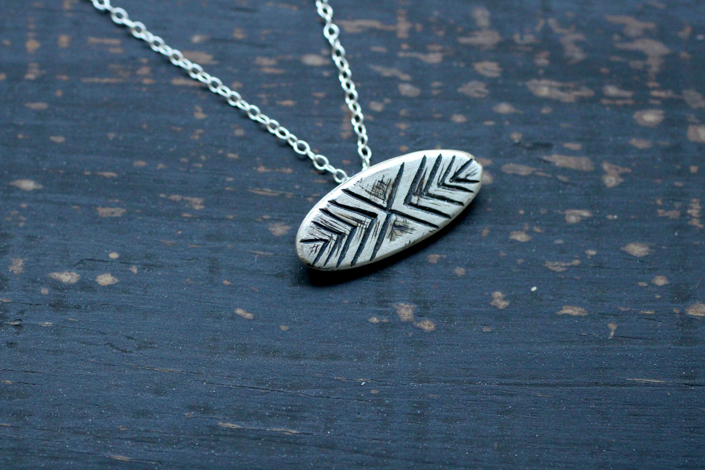 Silver necklace with a silver oval shaped pendant with a line pattern engraved with oxidation finish on a light background