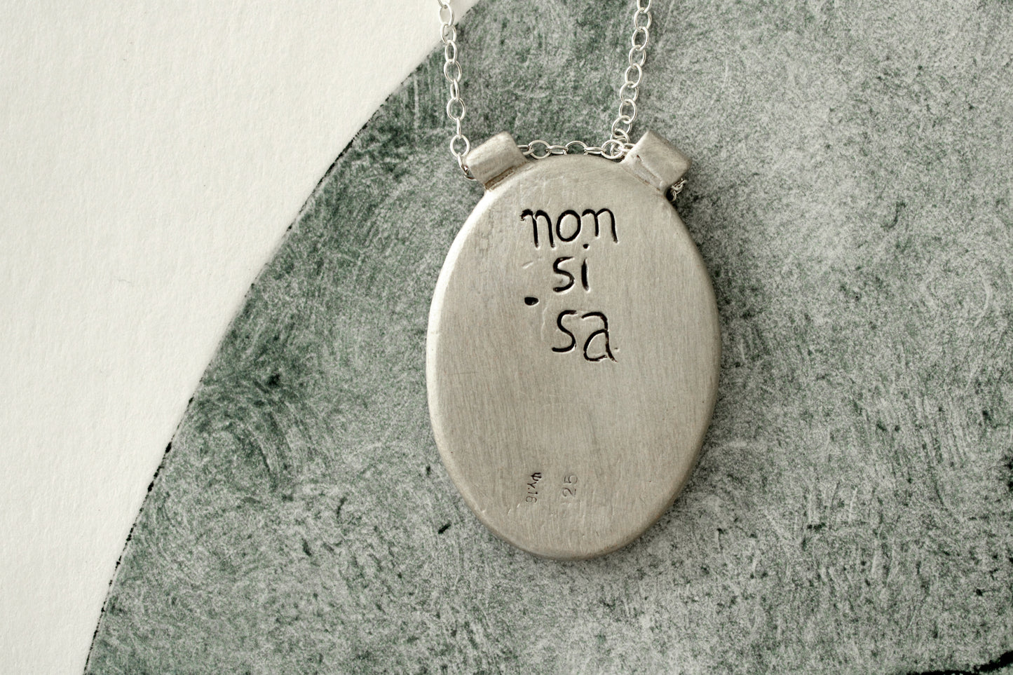 Silver necklace with a circular pendant with the brand name engraved in the back on a textured surface.