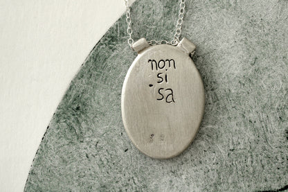 Silver necklace with a circular pendant with the brand name engraved in the back on a textured surface.