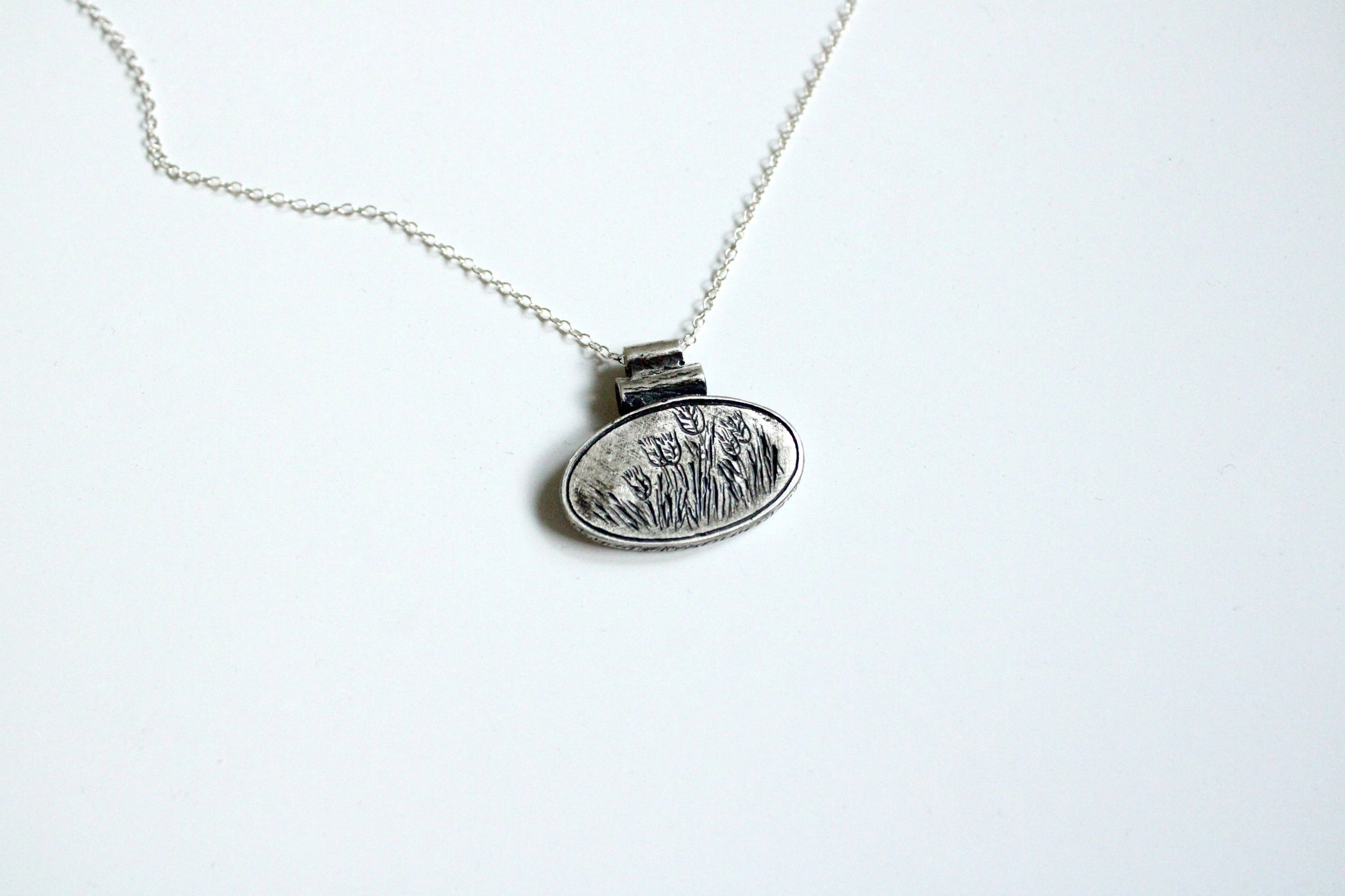 SIlver pendant with a silver chain with an oval shape with grain design engraved on a white background