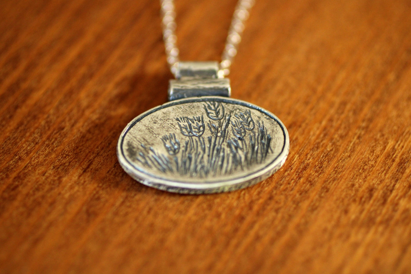 SIlver pendant with a silver chain with an oval shape with grain design engraved on a wooden background