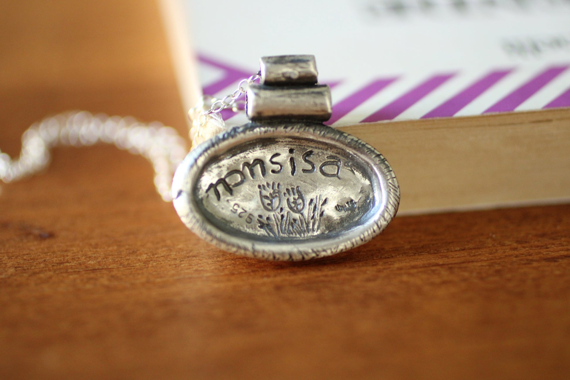 SIlver pendant with a silver chain with an oval shape with the brand name and grain design engraved  on the back on a wooden background