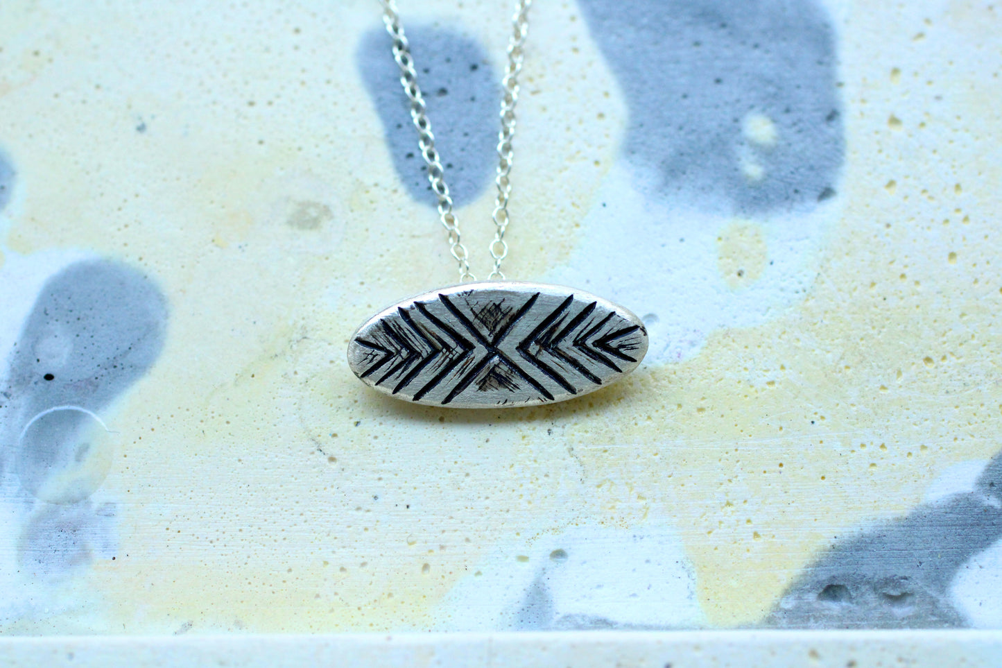 Silver necklace with a silver oval shaped  pendant with a line pattern engraved with oxidation finish on a light background