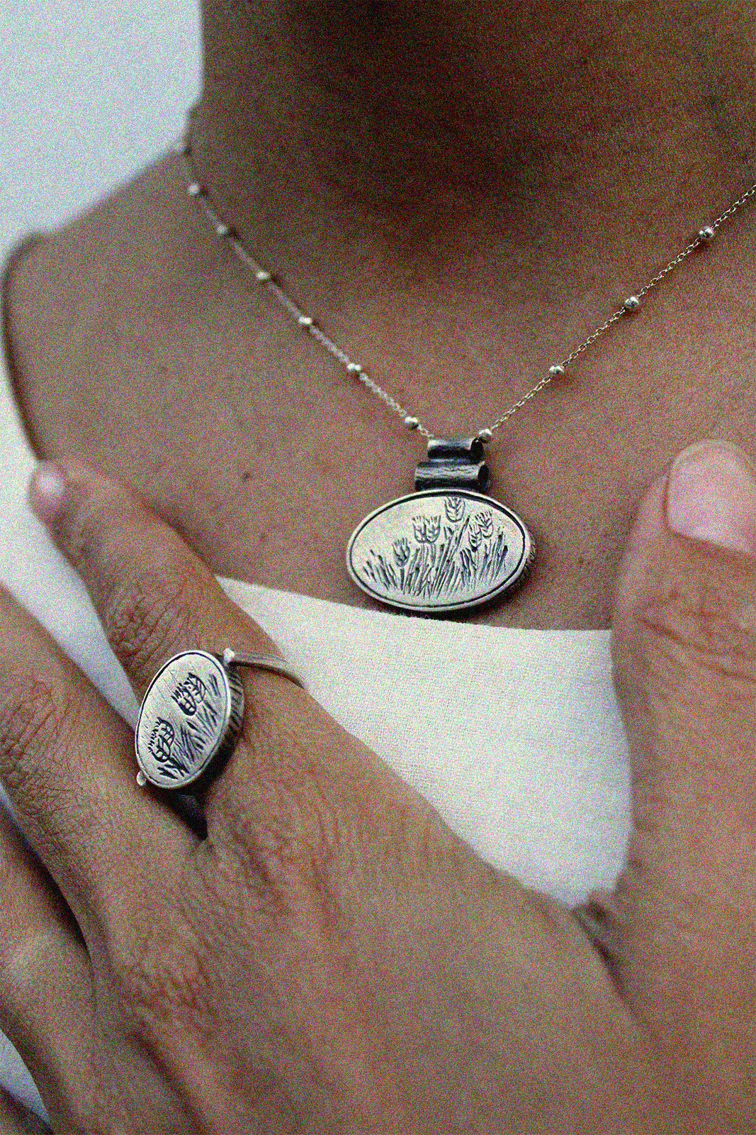 Close-up of a person wearing a silver necklace and ring with an engraved desig of wheat plants