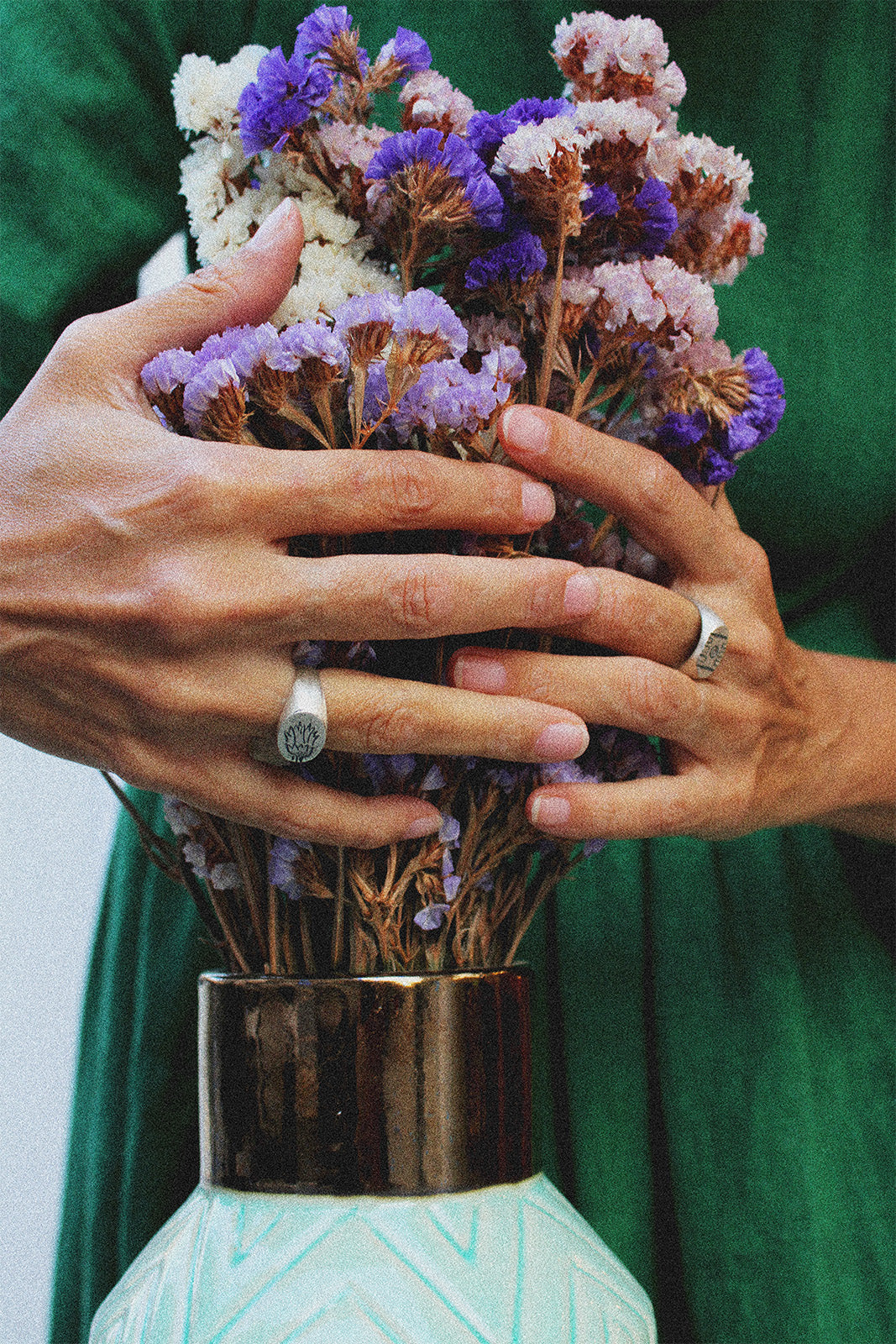 Person holding a vase with flowers against a green dress and wearing 2 silver rings 